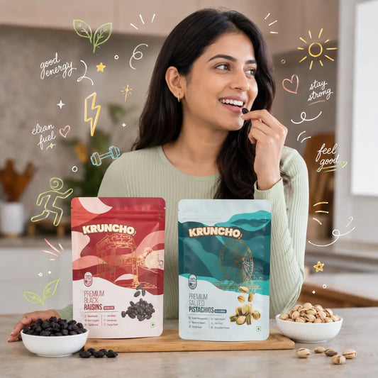 A smiling woman snacking on Kruncho Premium Black Raisins in a kitchen, next to packages of raisins and salted pistachios.