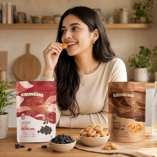 Lifestyle shot of a woman snacking on Kruncho dried apricots in a bright kitchen, with packages and bowls of Kruncho Black Raisins and Apricots displayed on the wooden counter.
