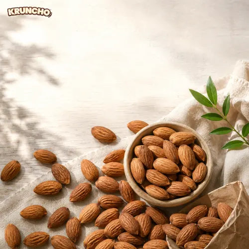 A small wooden bowl and paper bag filled with fresh raw almonds scattered on a beige cloth with the Kruncho logo.