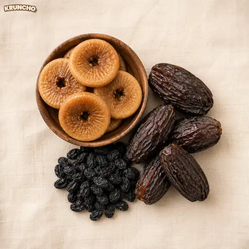 A wooden bowl of anjeer next to black raisins and large dates on a beige cloth.