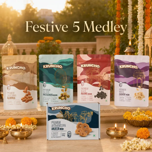 A festive outdoor setup featuring the Kruncho "Festive 5 Medley" collection, showcasing four dry fruit pouches and a boxed Anjeer pack surrounded by marigold flowers and lit brass diyas.