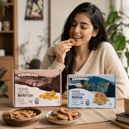 A smiling woman sitting at a table enjoys a dried fig, with KRUNCHO Premium Walnuts and Afghanistan Anjeer boxes and bowls of snacks displayed in front of her.