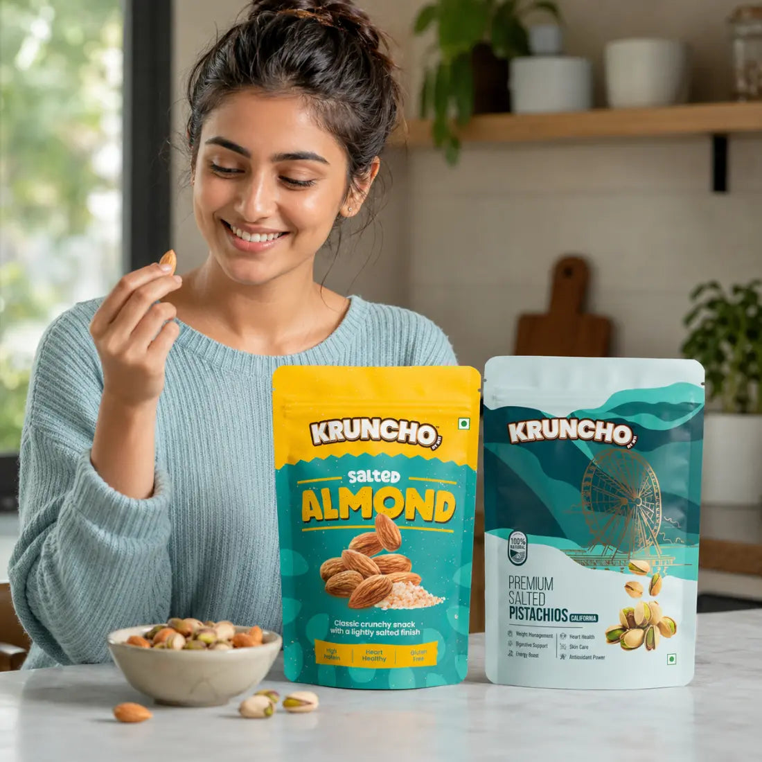 A woman enjoying a healthy snack at home, featuring Kruncho premium dry fruits including Salted Almonds and California Salted Pistachios packs on a kitchen counter.