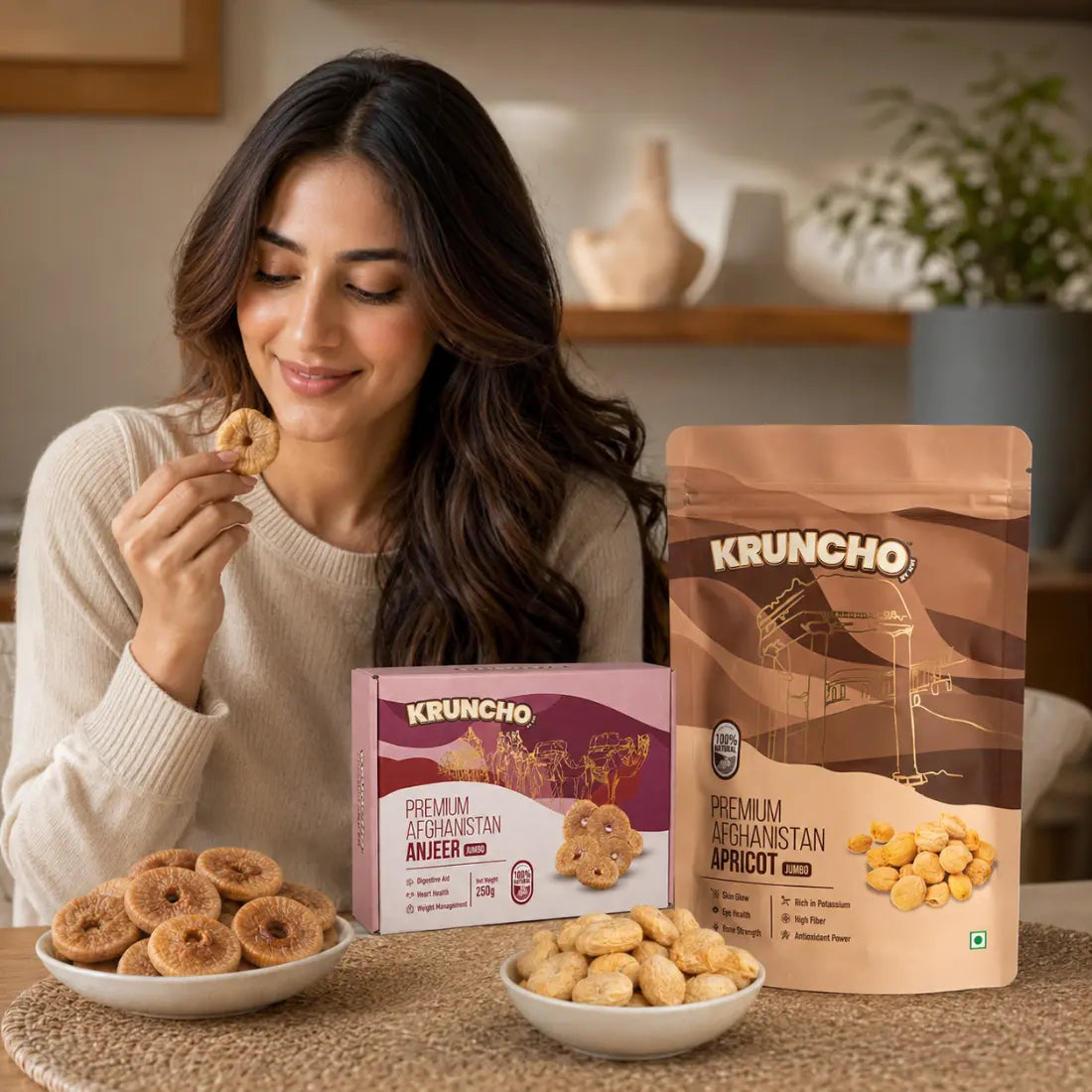 A woman smiling while holding a dried fig, with bowls of figs and apricots next to Kruncho Premium Afghanistan Anjeer and Apricot packaging.