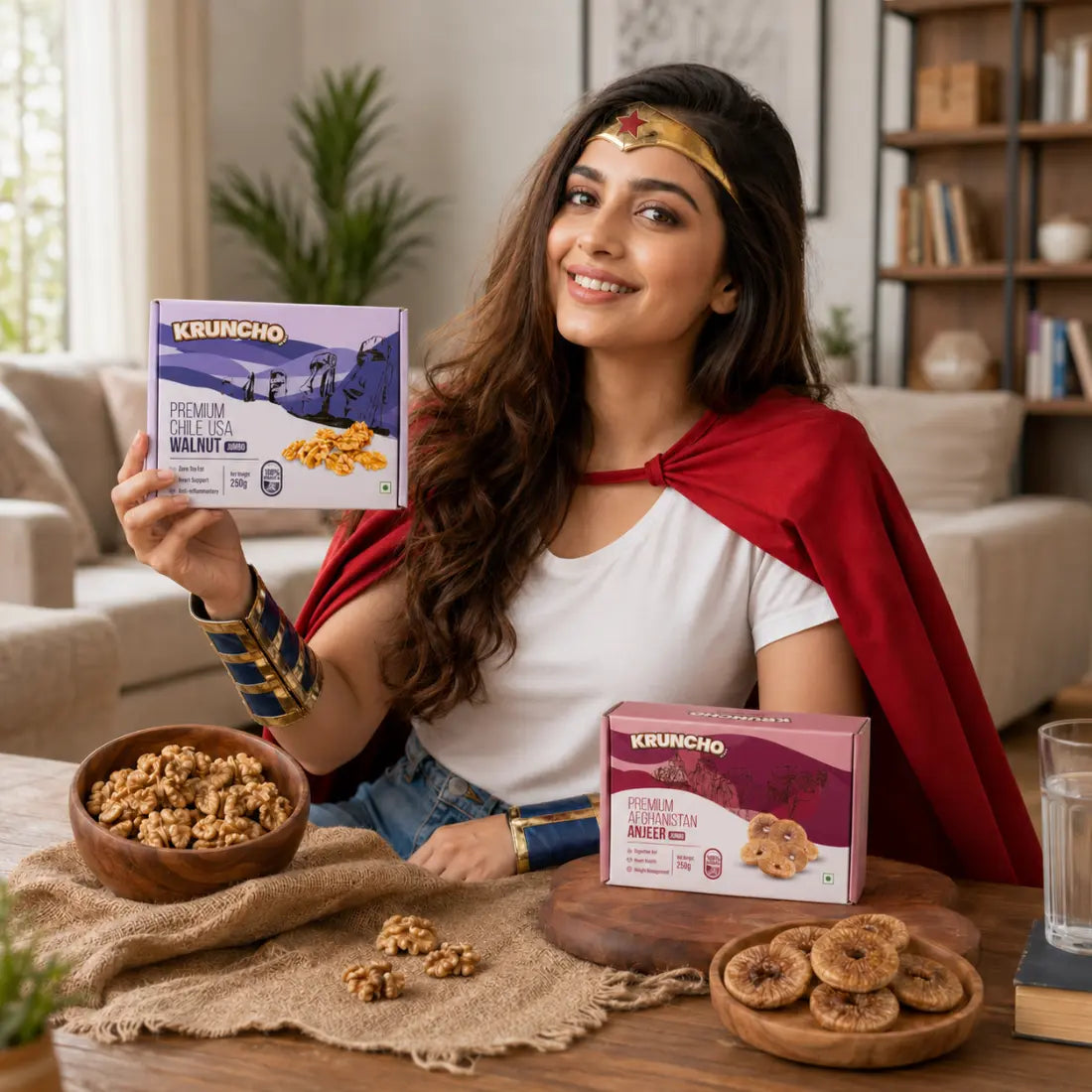 A smiling woman in a superhero cape and tiara holding a box of Kruncho Premium Chile USA Jumbo Walnuts at a wooden table featuring bowls of fresh walnuts and Kruncho Afghanistan Anjeer.